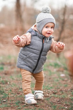 Bir yaşındaki bir çocuk pistte ilk adımlarını atıyor. Gri bir yelek ve bej pantolon giymiş seçici bir odak noktası var.