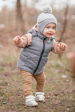 Bir yaşındaki bir çocuk pistte ilk adımlarını atıyor. Gri bir yelek ve bej pantolon giymiş seçici bir odak noktası var.