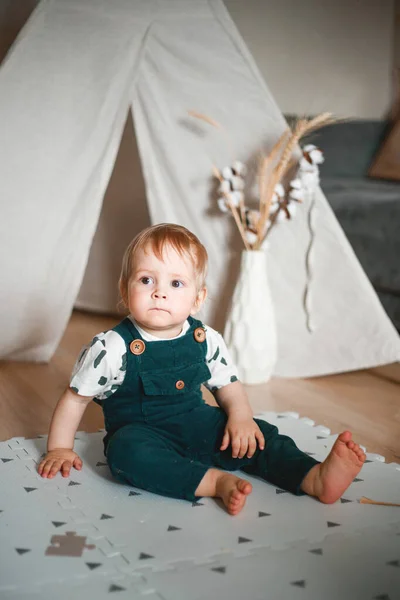 The baby is playing in his room in a wigwam, dressed in a green jumpsuit.