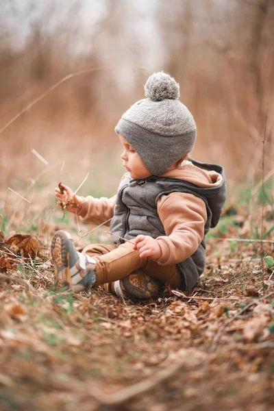 Küçük bir çocuk, gri bir jöle içinde bir orman yolunda oturuyor ve doğaya merakla bakıyor.