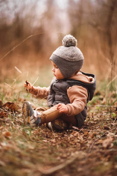 Küçük bir çocuk, gri bir jöle içinde bir orman yolunda oturuyor ve doğaya merakla bakıyor.