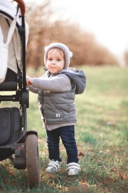Gri, ılık bir yelek ve gri şapkalı bir çocuk baharın arka planına karşı açık havada oyunlar oynuyor..