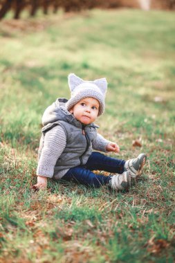 Gri, ılık bir yelek ve gri şapkalı bir çocuk baharın arka planına karşı açık havada oyunlar oynuyor..