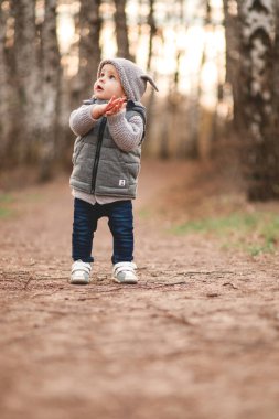 Gri, ılık bir yelek ve gri şapkalı bir çocuk baharın arka planına karşı açık havada oyunlar oynuyor..