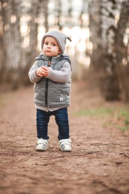 Gri, ılık bir yelek ve gri şapkalı bir çocuk baharın arka planına karşı açık havada oyunlar oynuyor..