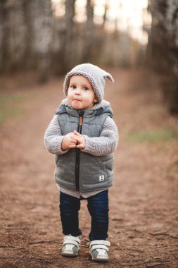 Gri, ılık bir yelek ve gri şapkalı bir çocuk baharın arka planına karşı açık havada oyunlar oynuyor..