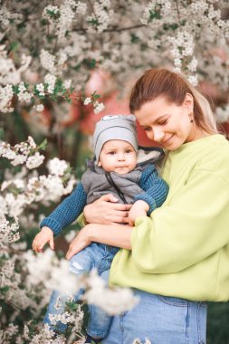 Bir anne, çiçek açan bir ağacın arka planında bir çocukla oynar ve konuşur. Mutlu gülümseyen insanlar.