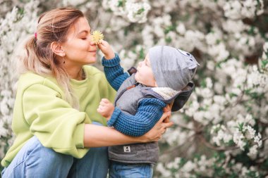 Bir anne, çiçek açan bir ağacın arka planında bir çocukla oynar ve konuşur. Mutlu gülümseyen insanlar.