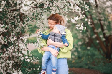 Bir anne, çiçek açan bir ağacın arka planında bir çocukla oynar ve konuşur. Mutlu gülümseyen insanlar.