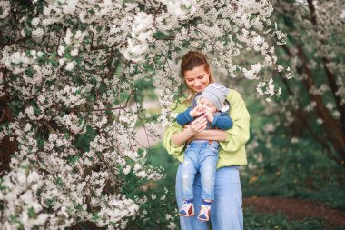 Bir anne, çiçek açan bir ağacın arka planında bir çocukla oynar ve konuşur. Mutlu gülümseyen insanlar.