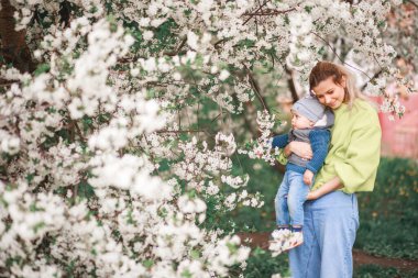 Bir anne, çiçek açan bir ağacın arka planında bir çocukla oynar ve konuşur. Mutlu gülümseyen insanlar.