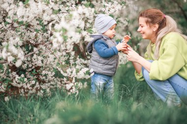 Bir anne, çiçek açan bir ağacın arka planında bir çocukla oynar ve konuşur. Mutlu gülümseyen insanlar.