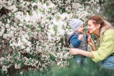Bir anne, çiçek açan bir ağacın arka planında bir çocukla oynar ve konuşur. Mutlu gülümseyen insanlar.