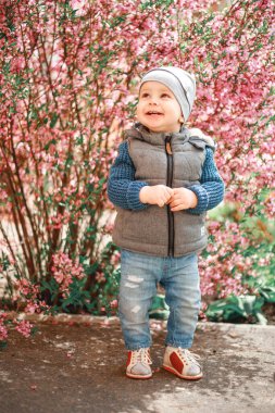 Kot giymiş bir çocuk İlkbaharda çiçekli bir çalılığın yanında duruyor