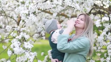 Güzel genç bir kadın kollarında sevimli bir çocuk tutuyor ve çiçek açan bir elma ağacının altında poz veriyor.