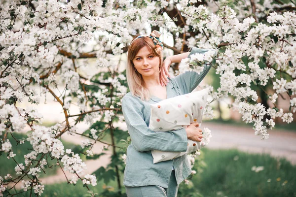 a girl hugs a large pillow with a casual expression on her face on a day off wears sleeping blue pajamas in a blooming Apple orchard in spring.