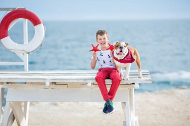 Yakışıklı çocuk teen happyly hayat şamandıra şamandıra kırmızı pantolon pantolon terlik ve t-shirt giyen yakın iki deniz yıldızları oynamaya holding deniz tarafında çocuk köpek onun arkadaş bulldog ile birlikte vakit geçirmek