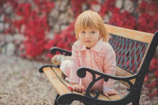 Baby cute girl with blond hair and pink apple cheek enjoying spring autumn time holiday posing in beautiful garden full of flowers on bench wearing pinky shirt dress pens — Stock Photo, Image