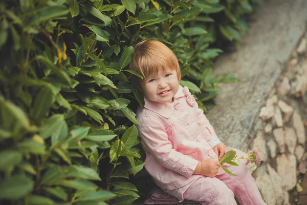 Bambino ragazza carina con i capelli biondi e guancia di mela rosa godendo di primavera autunno vacanza tempo in posa nel bellissimo giardino pieno di fiori che indossano penne abito camicia rosa — Foto Stock