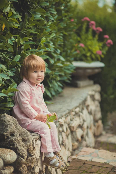 Bambino ragazza carina con i capelli biondi e guancia di mela rosa godendo di primavera autunno vacanza tempo in posa nel bellissimo giardino pieno di fiori che indossano penne abito camicia rosa — Foto Stock