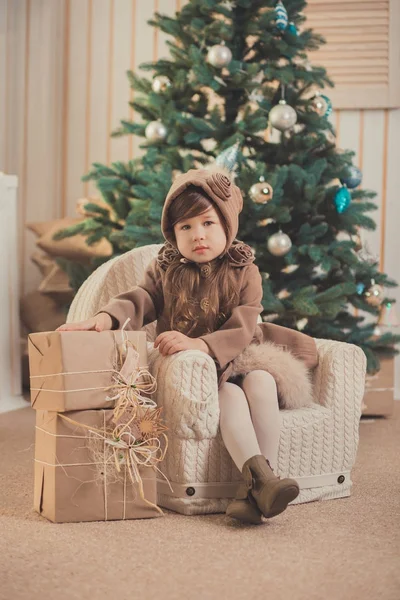 Νέοι μελαχρινή κουκλίτσα κυρία κορίτσι κομψά ντυμένος άνετα ζεστό χειμώνα γκρι μπουφάν με γούνα θέτοντας συνεδρίαση στέκεται στο studio κοντά στο δέντρο Χριστούγεννα Πρωτοχρονιά και παρουσιάζει. — Φωτογραφία Αρχείου