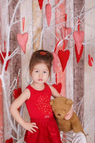 Joven morena dolly lady girl elegante vestido con zapatos de correa de vestido rojo sonriendo posando sentado en el estudio en una silla blanca con oso de peluche y árbol con corazones con labios de puchero y mejillas rosas . —  Fotos de Stock