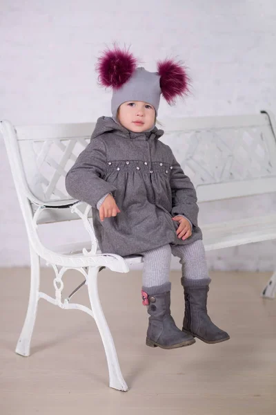 Bonita chica con cabello moreno elegante vestida con chaqueta gris y cálido sombrero de invierno alegremente sonriente posando a la cámara para la sesión de fotos en el moderno estudio de sala de exposición con botas de bebé . — Foto de Stock