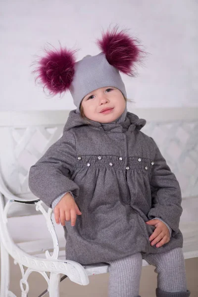 Bonita chica con cabello moreno elegante vestida con chaqueta gris y cálido sombrero de invierno alegremente sonriente posando a la cámara para la sesión de fotos en el moderno estudio de sala de exposición con botas de bebé . — Foto de Stock