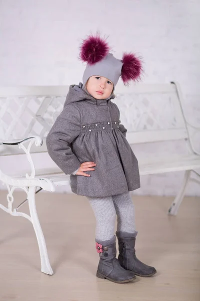 Hübsches Mädchen mit brünetten Haaren stilvoll gekleidet in grauer Jacke und warmem Wintermütze glücklich lächelnd posiert für Fotosession in modernen Showroom-Studio mit Babystiefeln. — Stockfoto