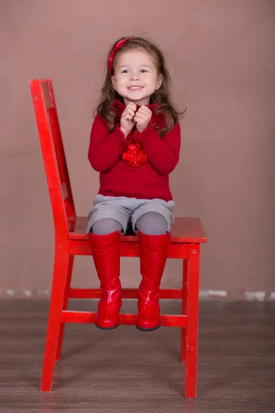 Menina bonita com cabelo morena elegante vestido camisola vermelha pulôver calções de inverno quentes calças alegremente sorrindo posando câmera fotossessão no estúdio showroom moderno vestindo botas de bebê sentado na cadeira . — Fotografia de Stock