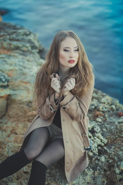 Seductora chica sexy con románticos labios rojos y mejillas meñique usando elegante vestido de chaqueta y pantalones cortos con botas de otoño posando en la playa rocosa junto al mar con maquillaje en la cara i episodio romántico — Foto de Stock