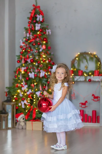 Menina bonita nova em azul branco elegante vestido de noite sentado no chão perto da árvore de Natal e apresenta em uma véspera de ano novo. Interior com decorações de Natal. DOF rasa e fundo bokeh — Fotografia de Stock