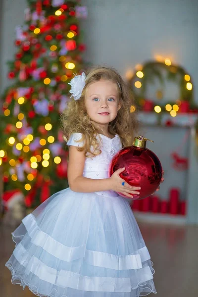 Menina bonita nova em azul branco elegante vestido de noite sentado no chão perto da árvore de Natal e apresenta em uma véspera de ano novo. Interior com decorações de Natal. DOF rasa e fundo bokeh — Fotografia de Stock