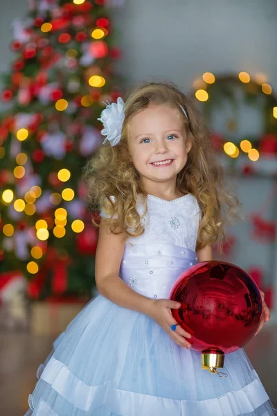 Menina bonita nova em azul branco elegante vestido de noite sentado no chão perto da árvore de Natal e apresenta em uma véspera de ano novo. Interior com decorações de Natal. DOF rasa e fundo bokeh — Fotografia de Stock