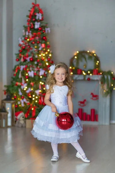 Menina bonita nova em azul branco elegante vestido de noite sentado no chão perto da árvore de Natal e apresenta em uma véspera de ano novo. Interior com decorações de Natal. DOF rasa e fundo bokeh — Fotografia de Stock