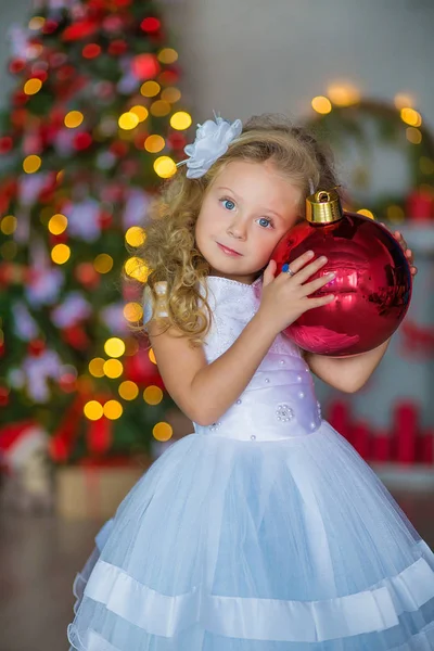 Menina bonita nova em azul branco elegante vestido de noite sentado no chão perto da árvore de Natal e apresenta em uma véspera de ano novo. Interior com decorações de Natal. DOF rasa e fundo bokeh — Fotografia de Stock