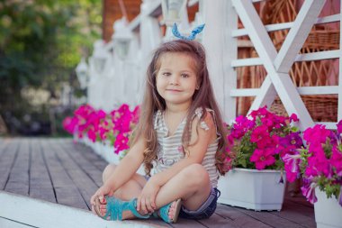 Stylish beautifull cute baby girl with brunette hair posing on wooden garden full of flowers wearing tiny jeans shirts and airy skivy underwaist and blue sandals clipart