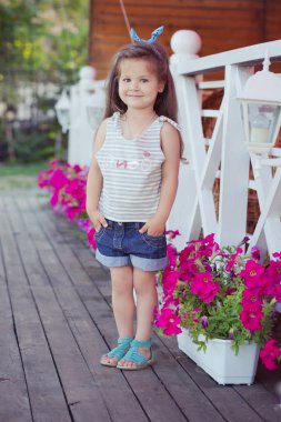 Esmer saçlı küçük kot gömlek ve havadar skivy underwaist ve mavi sandalet giyen çiçek ahşap Bahçe dolu poz şık güzel şirin bebek kız