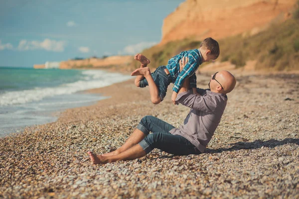 Wzruszające sceny atrakcyjne ojca i syna cieszyć się letnie wakacje razem grając na kamienistej plaży stylowe koszulkę i moda niebieski jeans zarówno boso z adore krajobraz na tle — Zdjęcie stockowe