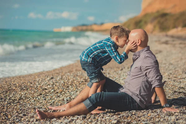 Emocionante escena atractiva de padre e hijo disfrutan de vacaciones de verano juntos jugando en la playa de piedra con camisa elegante y vaqueros azules de moda, ambos descalzos con adoro paisaje en el fondo — Foto de Stock