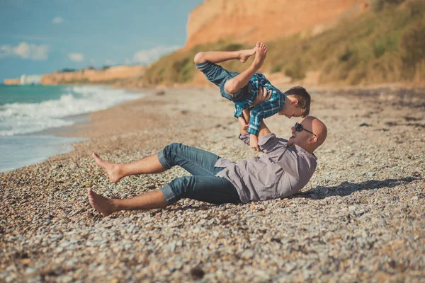 Emocionante escena atractiva de padre e hijo disfrutan de vacaciones de verano juntos jugando en la playa de piedra con camisa elegante y vaqueros azules de moda, ambos descalzos con adoro paisaje en el fondo — Foto de Stock