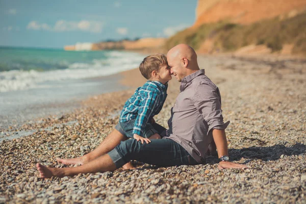 Emocionante escena atractiva de padre e hijo disfrutan de vacaciones de verano juntos jugando en la playa de piedra con camisa elegante y vaqueros azules de moda, ambos descalzos con adoro paisaje en el fondo — Foto de Stock