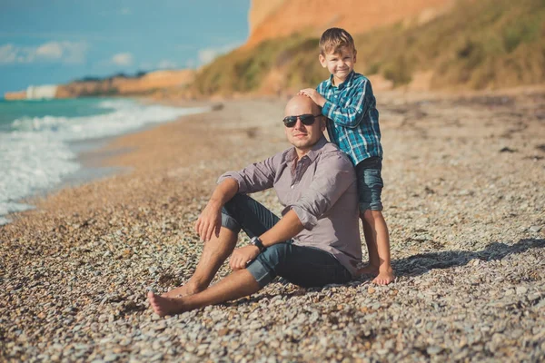 Emocionante escena atractiva de padre e hijo disfrutan de vacaciones de verano juntos jugando en la playa de piedra con camisa elegante y vaqueros azules de moda, ambos descalzos con adoro paisaje en el fondo — Foto de Stock