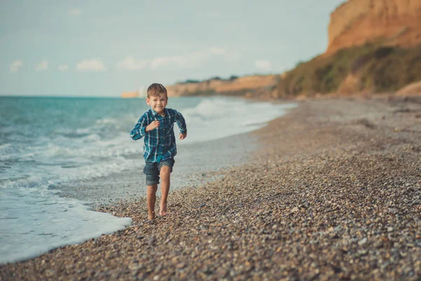 Симпатичний хлопчик дитина в стильній сорочці і блакитних джинсах босоніж позує біжить на кам'яному пляжі з розкішним океанським морем пейзаж піщаний скелястий мис — стокове фото
