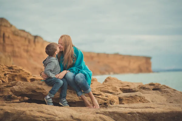 Escena encantadora familiar de la hermosa madre posando besando a su lindo hijo guapo en la playa junto al mar durante las vacaciones de verano en el resort oceánico con arena acantilado paisaje — Foto de Stock