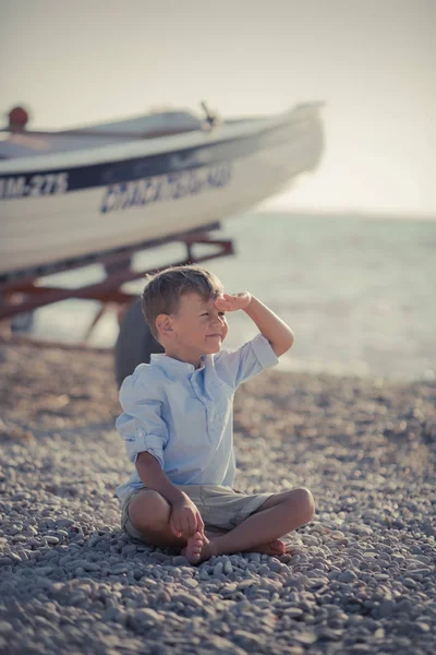Гарний красивий хлопчик на пляжі поруч з дерев'яним човном у вишуканій стильній блакитній сорочці та шортах з шибеницями насолоджується літнім часом наодинці на чудовому океані — стокове фото