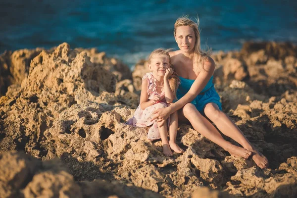 Moeder van de schattige dame zittend op rotsen strand in lichte blauwe jurk samen met haar schattige blonde dochtertje draagt roze luchtige jurk genieten van tijd weekend samen familie en zee oceaan. — Stockfoto