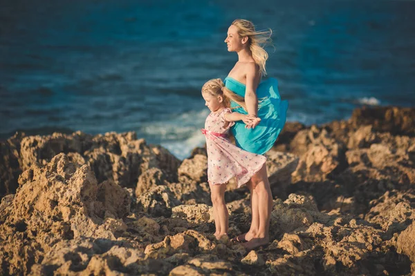 Adorable lady mother sitting on rocks beach in light blue dress together with her cute little blond daughter wearing pink airy dress enjoying time weekend together family and sea ocean.