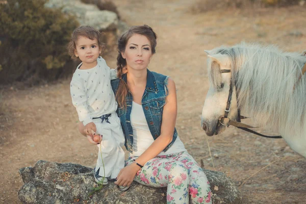 Romantisch tafereel mooie dame jonge moeder met haar schattige baby dochter samen genieten van tijd in de stad dorp park wandelen voeding wit paard pony — Stockfoto