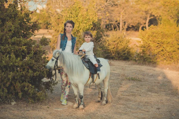 Cena romântica bela senhora jovem mãe com sua filha bebê bonito desfrutar de tempo juntos no parque da aldeia da cidade andando alimentando cavalo branco pônei — Fotografia de Stock